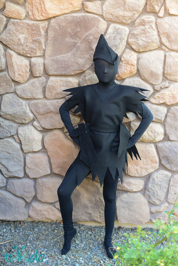 Young girl wearing an all black Peter Pan's escaped shadow costume in front of a stone wall. 
