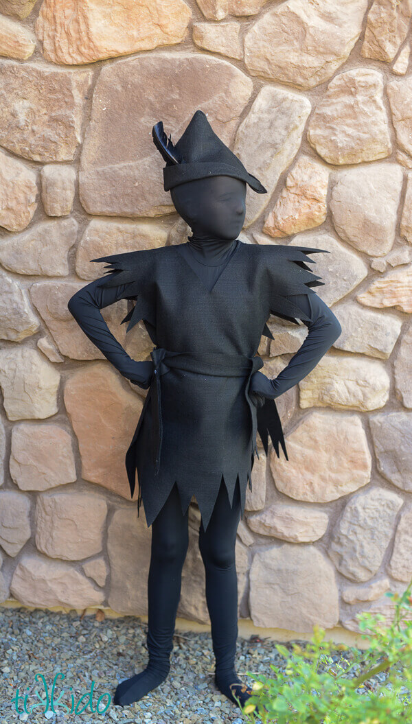 Young girl wearing an all black Peter Pan shadow costume in front of a stone wall. 