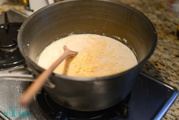 Shredded white cheddar cheese being added to roux and beer mixture in a saucepan, being stirred with a wooden spoon