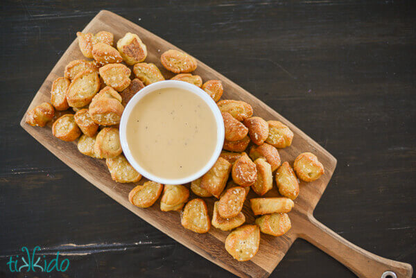 Overhead view of a wooden cutting board heaped with homemade pretzel bites, with a white bowl of beer cheddar dipping sauce in the center.