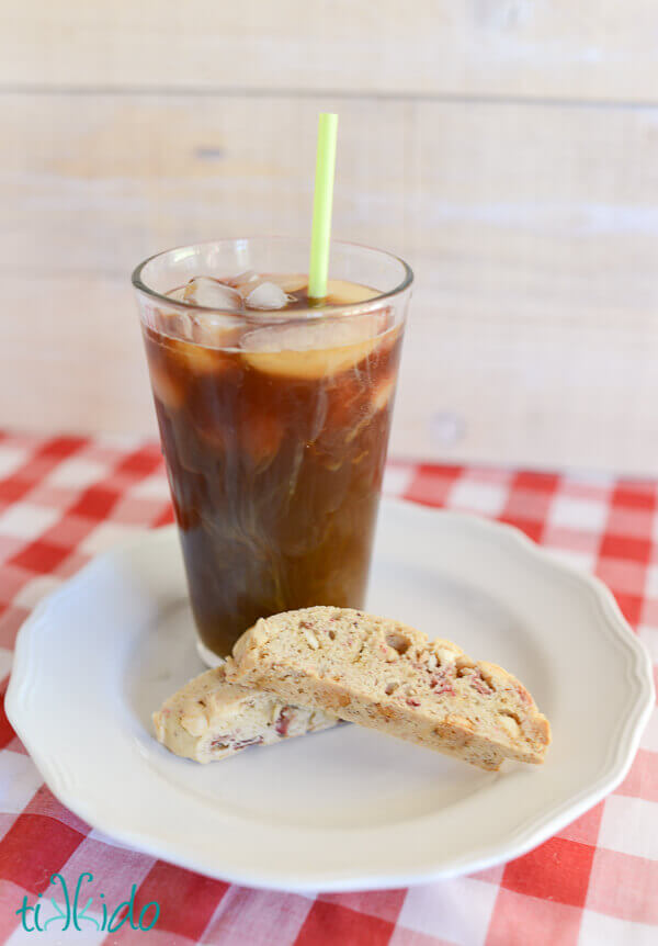 Two White chocolate strawberry biscotti cookies and an iced coffee on a white plate, sitting on a red and white checkered tablecloth.