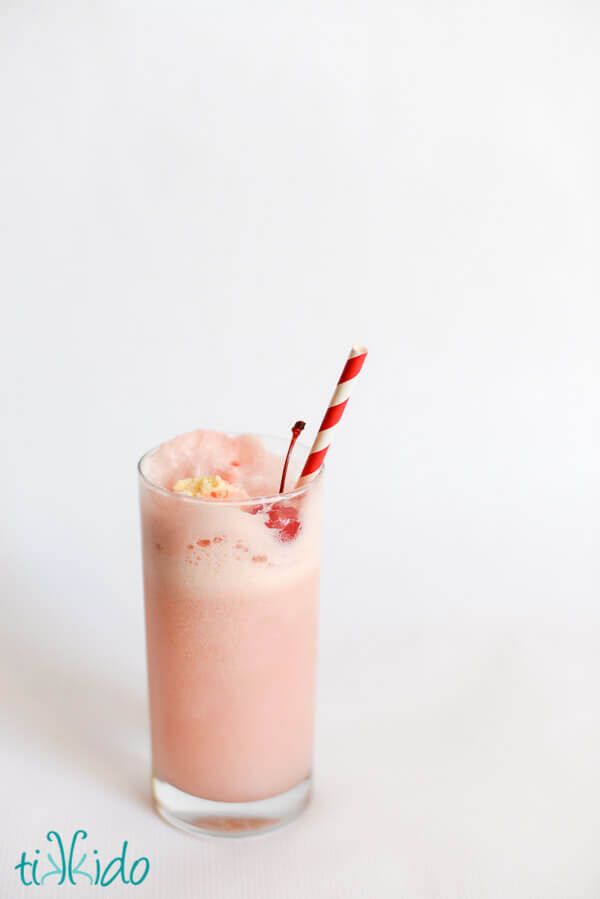 Shirley Temple Ice Cream Float topped with a maraschino cherry and a red and white striped paper straw, on a white background.