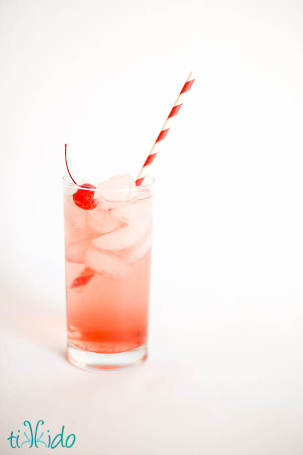 Shirley temple in a tall glass with a red and white striped straw on a white background.