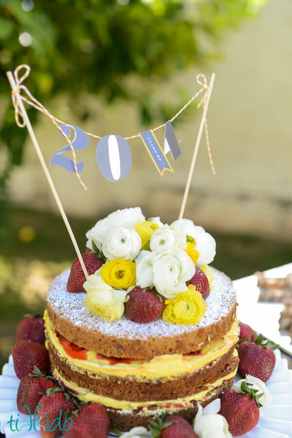 Graduation cake topper on a naked cake decorated with strawberries and fresh flowers.