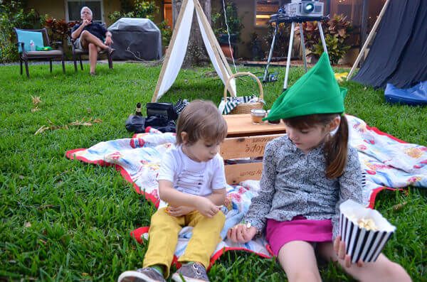 Little boy and little girl sitting on a picnic blanket in a backyard, eating popcorn and wearing a green peter pan hat.