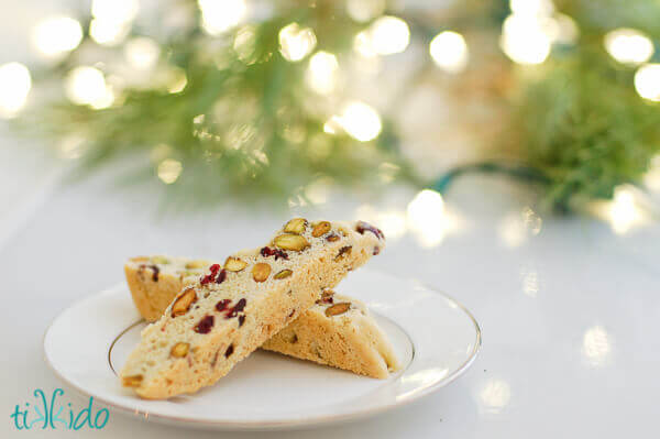 Two slices of Cranberry Pistachio Biscotti on a white plate in front of Christmas lights and fresh greens.