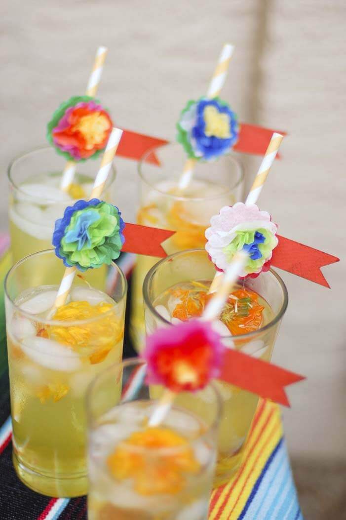 Light yellow cocktail in a tall glass with a striped paper straw embellished with a miniature Mexican style tissue paper flower.