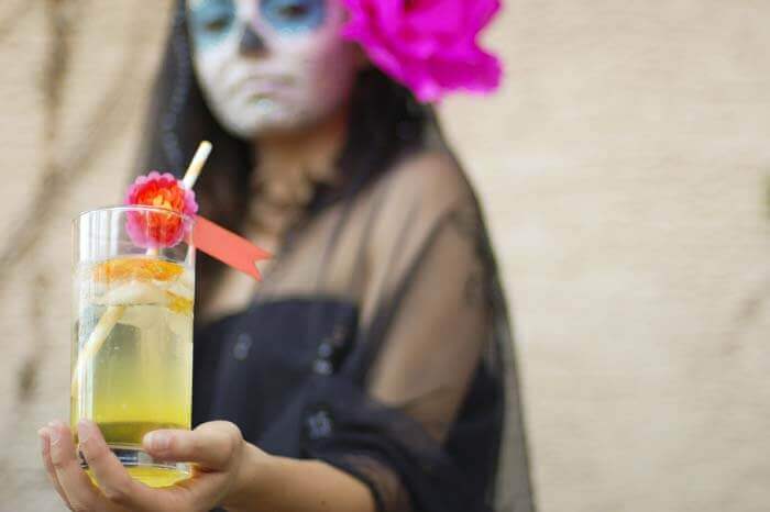 Woman with dia de los muertos face paint, a large pink paper flower in her hair, and a black veil holding a Marigold Muerte cocktail.