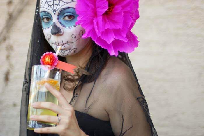 Woman with dia de los muertos face paint, a large pink paper flower in her hair, and a black veil sipping a Marigold Muerte cocktail.