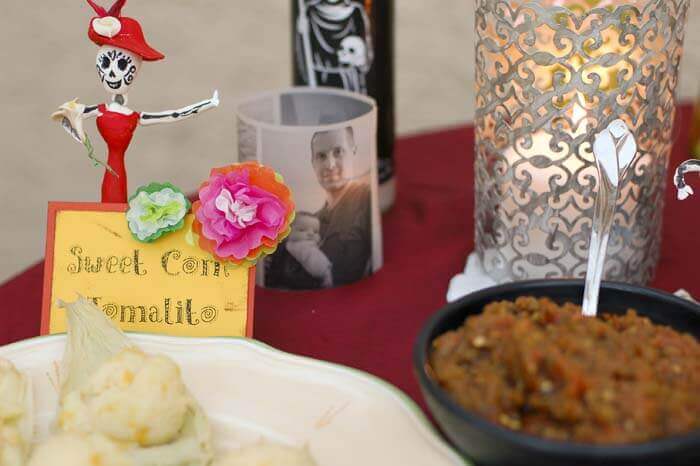 Candles, day of the dead figurines, and a card with miniature tissue paper flowers labeled "Sweet Corn Tomalito" next to plate of microwave sweet corn pudding.