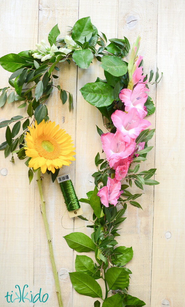 Fresh flowers, fresh greens, and green floral wire  being bound into a fresh flower garland on a whitewashed wooden backdrop.