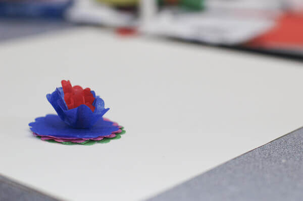 Scalloped circles of colorful tissue paper being glued together and fluffed into a miniature tissue paper flower.