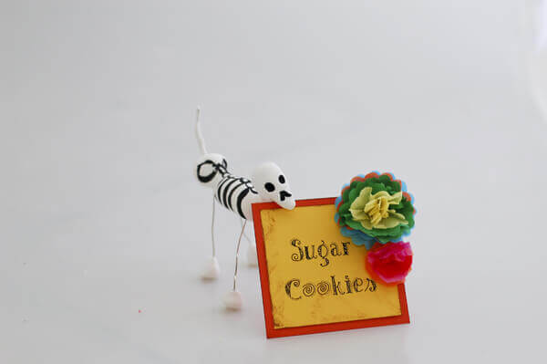 Colorful Mexican style miniature tissue paper flowers on a table tent being held up by a Dia de los Muertos skeletal dog sculpture.