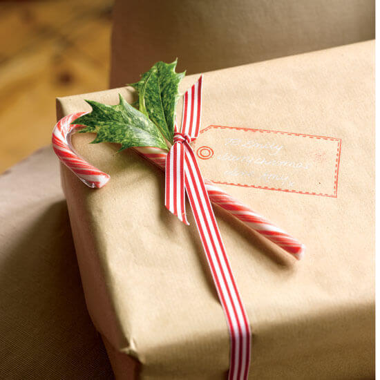 Package wrapped in kraft paper with a stamped gift tag, tied with a red and white striped ribbon, and embellished with holly leaves and a candy cane.