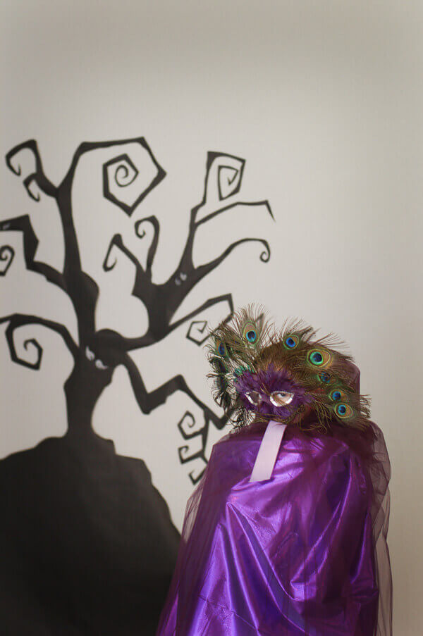 Young girl wearing a DIY purple tulle cape and masquerade mask in front of a spooky tree background.