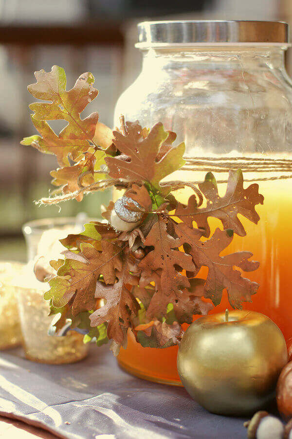 Felt acorns, twine, and oak leaves decorating a drink dispenser full of apple cider.