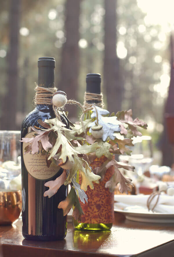 Felt Acorns and oak leaves decorating two wine bottles sitting on a table outside.