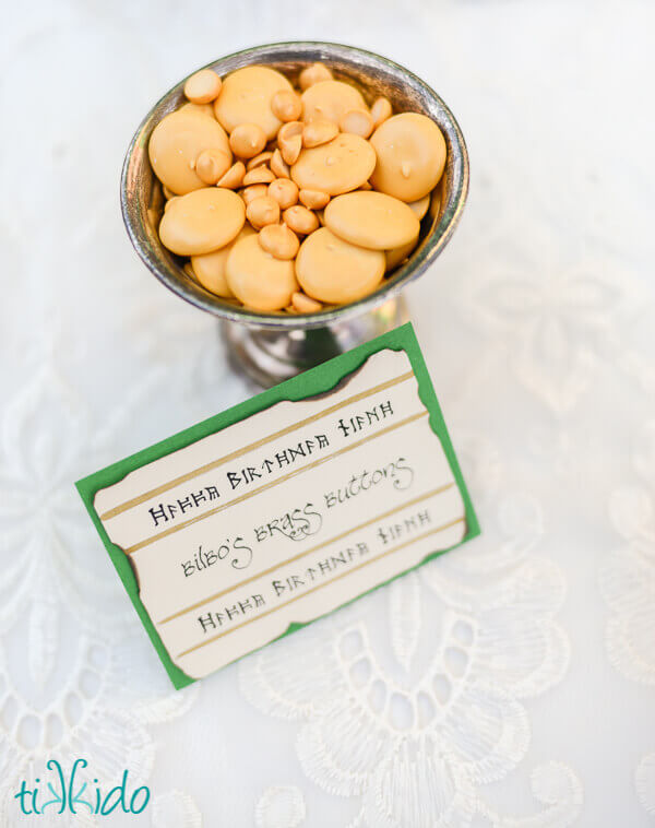 Disks of dried royal icing painted a golden color and labeled Bilbo's Brass Buttons in a small metal dish.