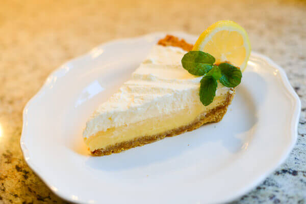 Slice of lemon pie topped with whipped cream and sitting on a white plate.  The pie is garnished with a slice of lemon and a sprig of mint.