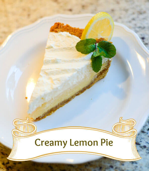 Slice of lemon custard pie on a graham cracker crust, topped with whipped cream, and garnished with fresh mint and a slice of lemon.
