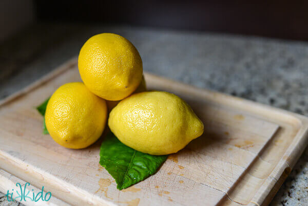 Four whole lemons stacked on a wooden cutting board.