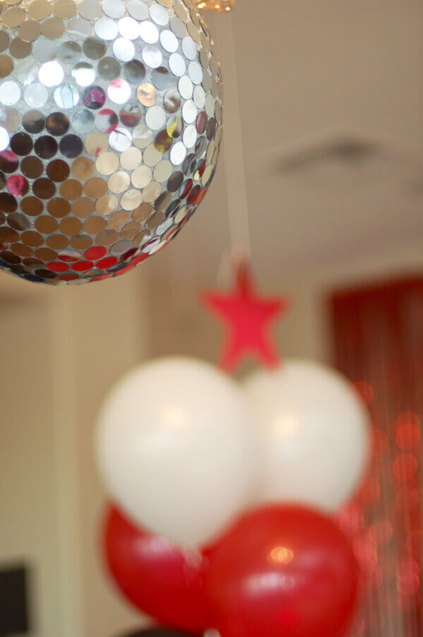 Piñata that looks like a disco ball hanging from the ceiling in front of red and white balloons.