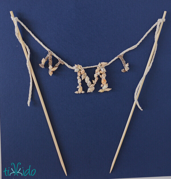 Nautical Monogram Cake Topper decorated with small shells and sand, on a dark blue background.