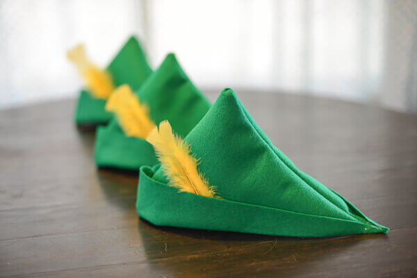 Three green felt peter pan hats with yellow feathers on a dark brown wooden table.