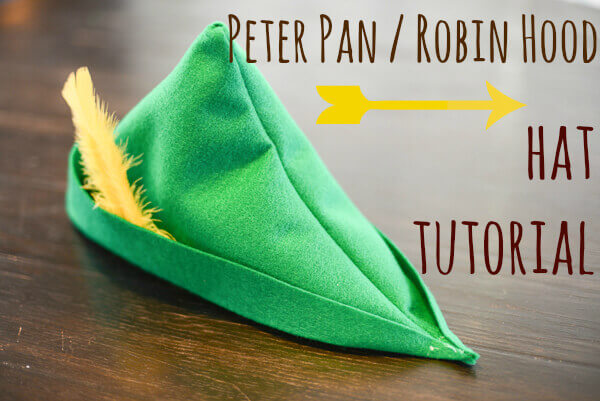 Green felt Peter Pan hat with a yellow feather tucked in the brim, on a dark brown wooden surface.