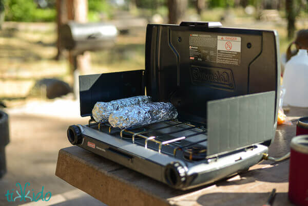 Corn wrapped in foil being cooked on a camp stove.