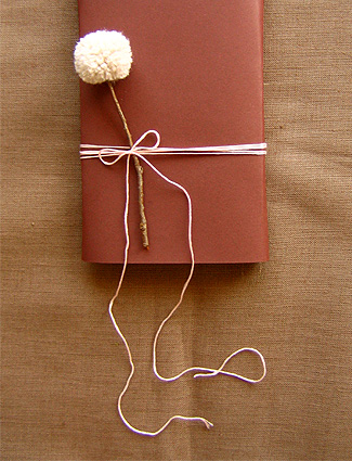 Package wrapped in reddish brown paper, tied with white string, and embellished with a yarn pom pom flower on a twig.