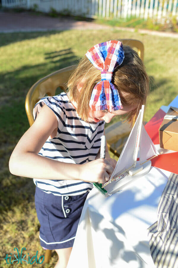 Little girl wearing a large fabric hair bow, striped shirt, and navy shorts, coloring a white toy sailboat.