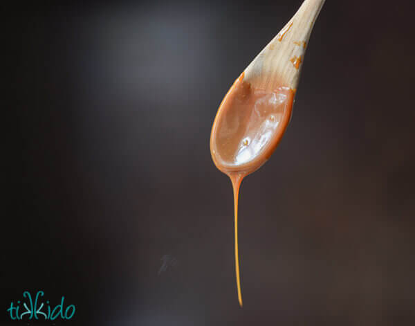Salted caramel sauce dripping off a wooden spoon against a dark brown background.