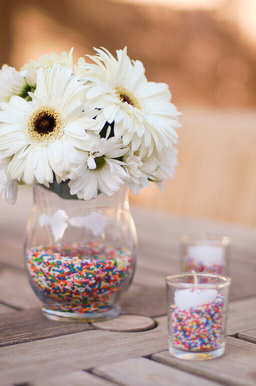White daisies in a vase filled with rainbow sprinkles.