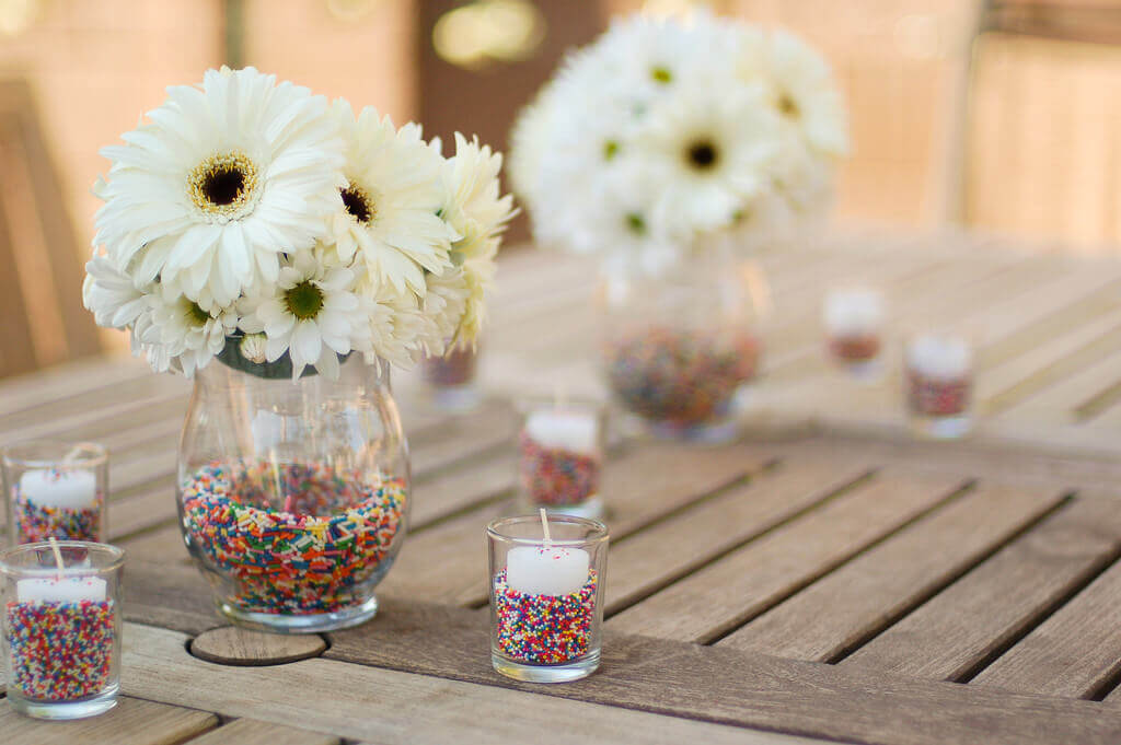 Rainbow sprinkle filled vases with white daisies, surrounded by votive candles surrounded by rainbow sprinkles.