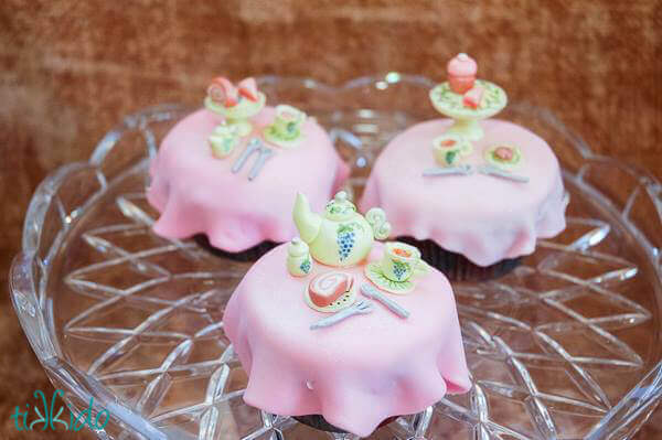 Three tea party cupcakes, featuring gum paste tea pots, cakes, and cups of tea, on a glass cake stand.