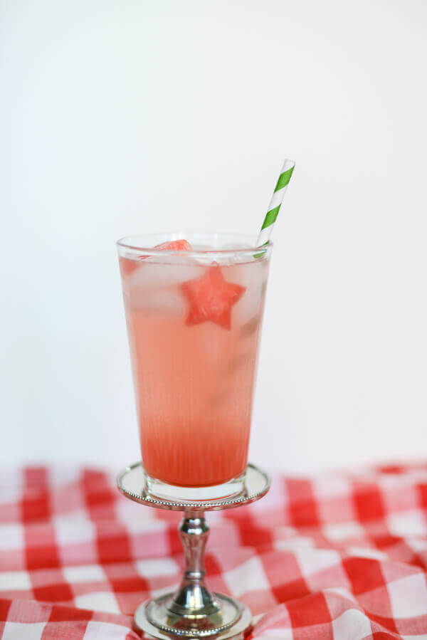 Pink watermelon drink with a star shaped piece of watermelon in a tall glass, with a green striped paper straw, on a white background.