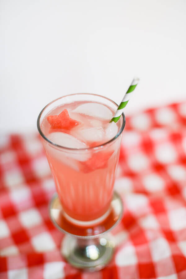 Pink watermelon drink with a star shaped piece of watermelon in a tall glass, with a green striped paper straw, on a red and white checkered background.