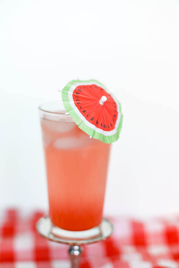 Pink watermelon drink in a tall glass with a watermelon themed cocktail umbrella.