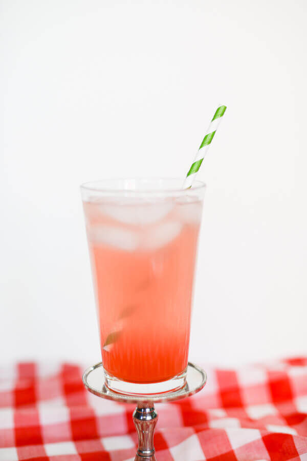 Pink watermelon drink in a tall glass with a green and white straw on a red and white checked tablecloth and white background.