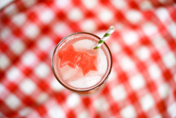 Top view of Watermelon Sparkler cocktail with two pieces of watermelon cut into star shapes in the drink, a green and white straw, on a red and white checked tablecloth.