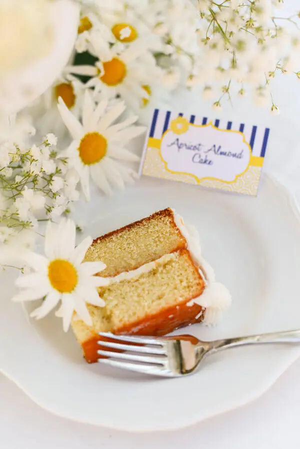 Slice of vanilla cake flavored with an amaretto cake soak and apricot glaze, with a sign reading "Apricot Almond Cake" in the background, next to flowers.