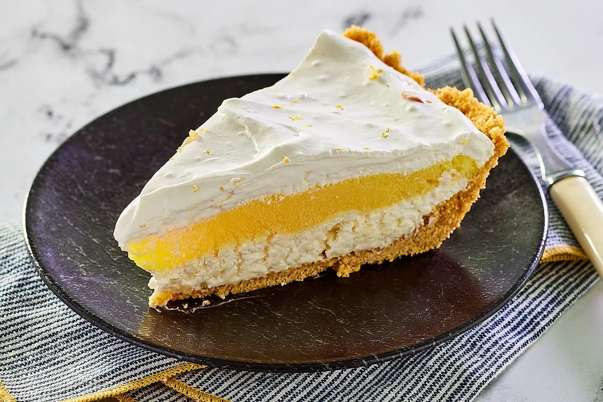 Piece of lemon lush pie with graham cracker crust on a black plate on a blue and white striped cloth.