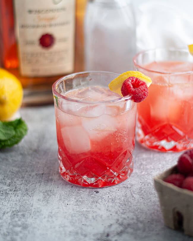 Deep pink raspberry bourbon smash in a cut crystal glass, garnished with a slice of lemon and a fresh raspberry.