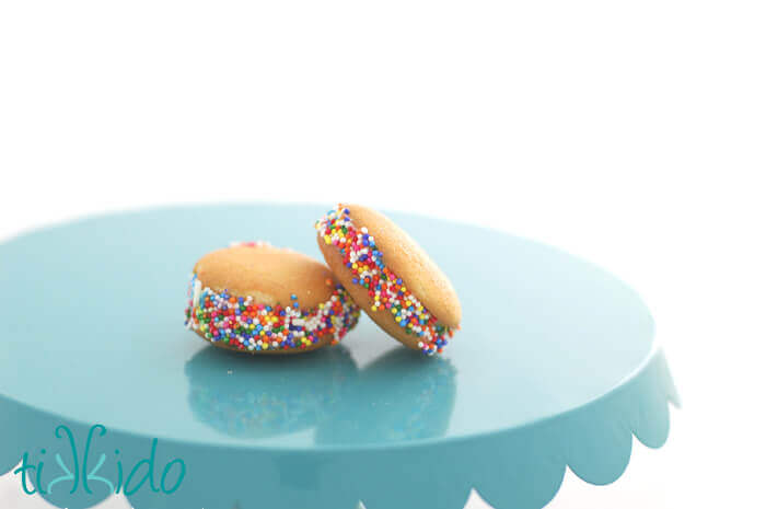 Two miniature Nilla Wafer ice cream sandwiches coated in rainbow sprinkles, on a blue cake stand.
