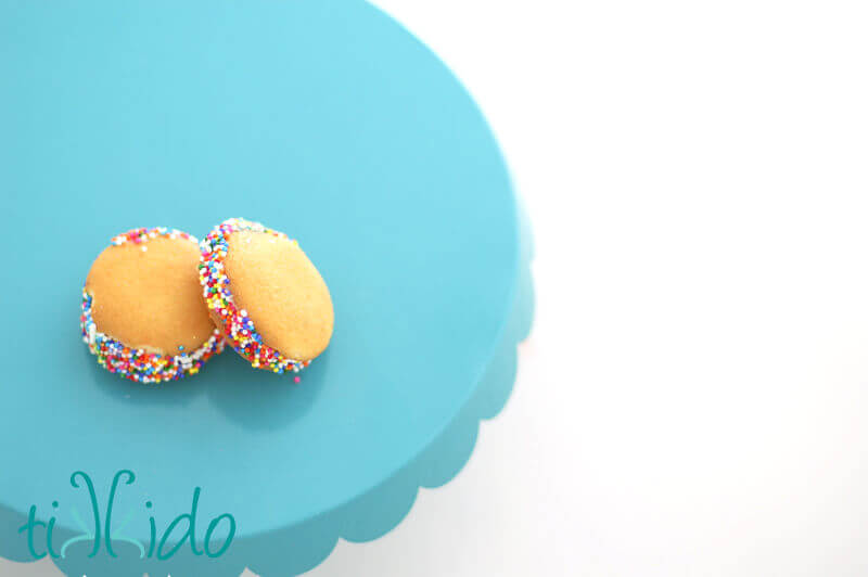 Two miniature Nilla Wafer ice cream sandwiches coated in rainbow sprinkles, on a blue cake stand.