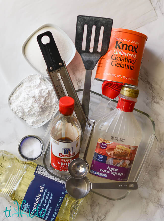 Ingredients for homemade marshmallow recipe including corn syrup, oil, gelatin, sugar, powdered sugar, salt, and vanilla, on a white marble background.