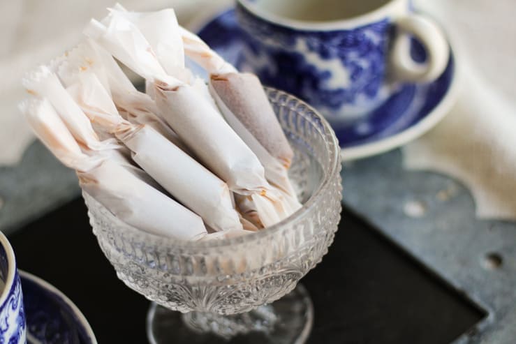 Bacon fat caramels wrapped in parchment paper in a footed glass bowl, in front of a blue and white tea cup.