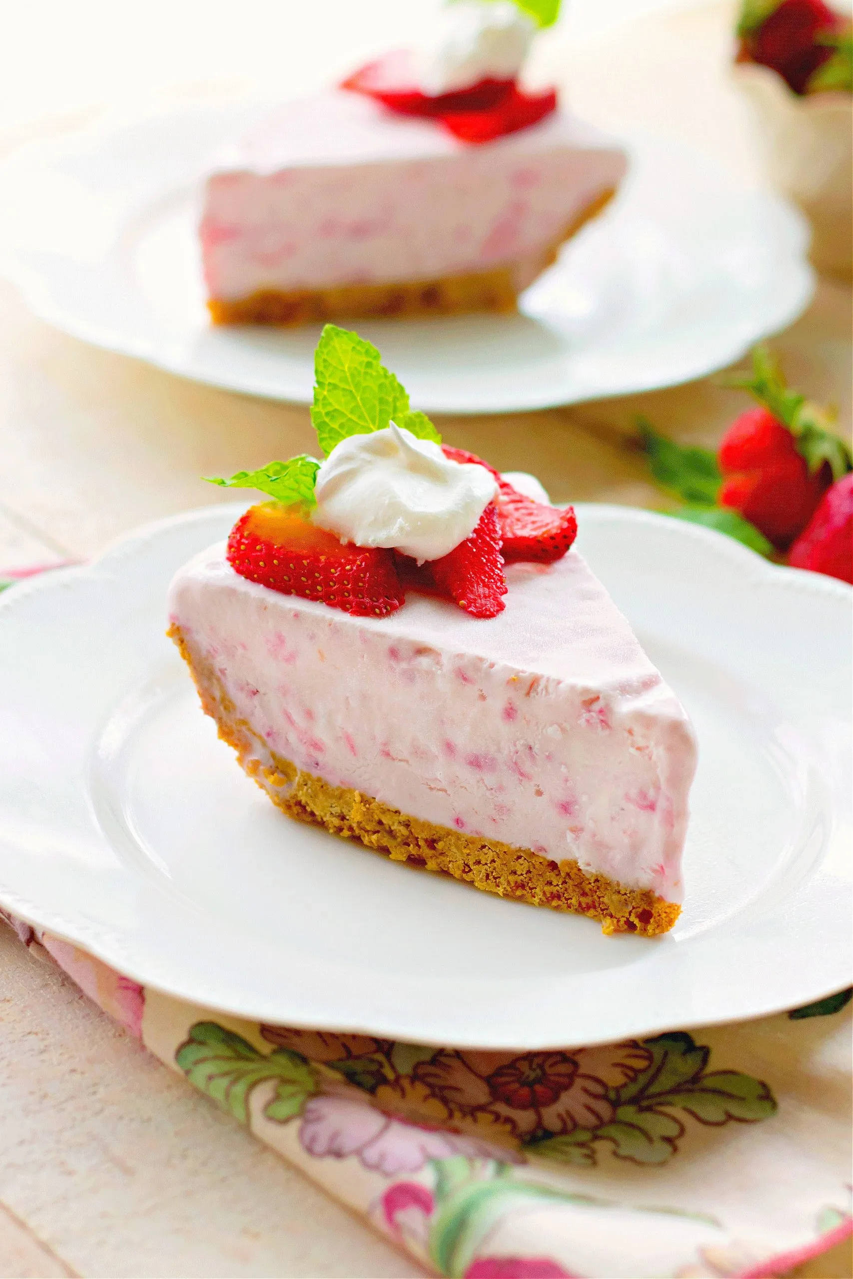 Slice of frozen strawberry cream pie with graham cracker crust on a white plate.  The slice of pie is garnished with slices of fresh strawberries and whipped cream.