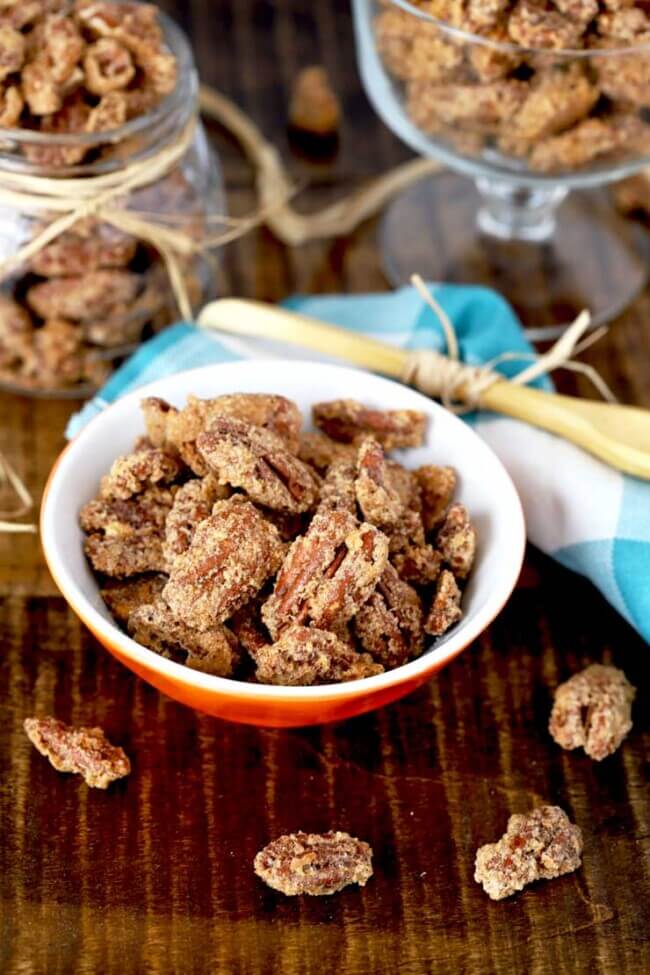 Three containers of candied pecans on a wooden surface next to a blue and white checked kitchen towel.